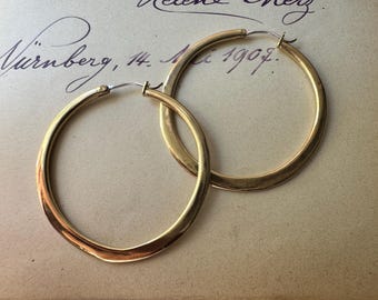May include: A pair of large, circular gold hoop earrings with a smooth, polished finish. The earrings feature a thin, metallic wire closure mechanism at the top, designed to secure the hoop in place when worn. The gold metal has a reflective surface.