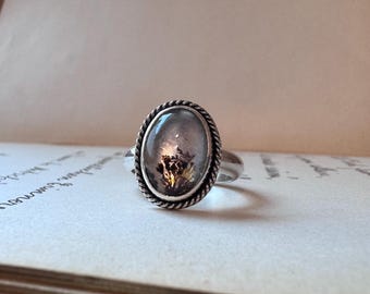 May include: A silver ring featuring an oval-shaped dendritic agate gemstone with dark, tree-like inclusions set within a twisted rope-style metal border. The band is smooth and polished, highlighting the contrast between the metallic setting and the stone.