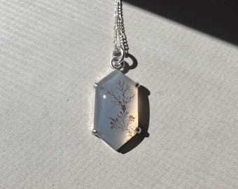 May include: A silver pendant necklace featuring a hexagonal, light blue gemstone with a dendritic pattern. The pendant is suspended from a delicate silver chain. The gemstone has a natural, organic appearance, with a tree-like design.