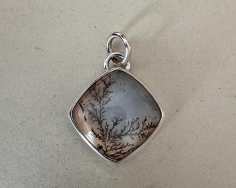 May include: A square-shaped pendant featuring a translucent stone with dark, branching, fern-like dendrite inclusions. The stone is encased in a polished silver metal setting with a circular bail at the top for attachment to a chain or cord.