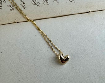 May include: A gold-toned necklace featuring a small, polished, three-dimensional heart pendant suspended from a delicate, fine-link chain. The metal surface of the heart is smooth and reflective, catching light to highlight its rounded, puffed shape.