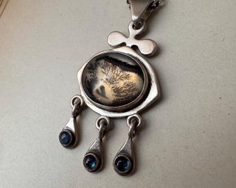 May include: A silver pendant necklace featuring a large oval dendritic agate stone with dark tree-like inclusions. The setting includes a decorative top piece and three dangling silver charms, each set with a small, round, dark blue gemstone.