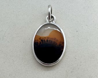 May include: An oval pendant set in a polished silver frame. The stone features a smooth, translucent surface with a distinct color gradient transitioning from a creamy white top to a warm amber middle and a solid, opaque black base with dark inclusions.
