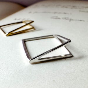 May include: Two geometric rings, one in a polished silver tone and one in a gold tone. Each ring features a square base with an angled, raised bar that connects to one corner, creating a three-dimensional, open-frame structure with clean lines.