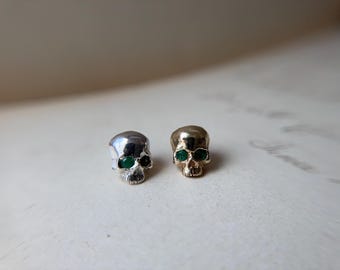 May include: Two small skull-shaped stud earrings. One earring is silver-toned with green gemstone eyes, and the other is gold-toned with green gemstone eyes. Both skulls feature detailed facial structures including eye sockets, nasal cavities, and teeth.