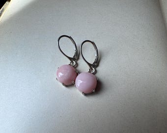 May include: A pair of lever-back earrings featuring round, pale pink gemstones. The gemstones are set in a silver-colored metal. The earrings are displayed on a light-colored surface.