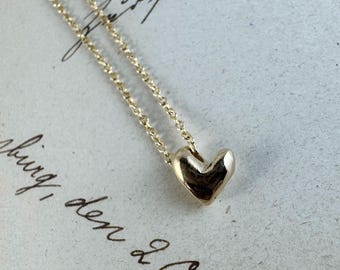 May include: A gold-colored necklace featuring a small, smooth, three-dimensional heart-shaped pendant suspended from a delicate, thin link chain. The metal surface has a polished, reflective finish that catches the light, creating subtle highlights.