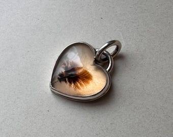 May include: A heart-shaped pendant set in a polished silver-toned metal frame. The center features a smooth, translucent stone with a natural, dark brown, fern-like dendritic inclusion that spreads across the cream-colored surface of the gem.