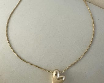 May include: A gold-toned necklace featuring a delicate, thin chain and a single, smooth, puffed heart-shaped pendant. The pendant has a polished, reflective surface that catches the light, suspended centrally from the fine, linked metal chain.