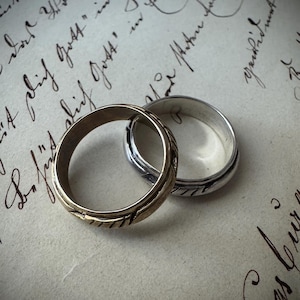 Può includere: Due fedi nuziali, una d'oro e una d'argento, poggiano su uno sfondo di carta vintage con scritte a mano. L'anello d'oro ha un design strutturato, mentre l'anello d'argento è liscio. Gli anelli sono intrecciati.