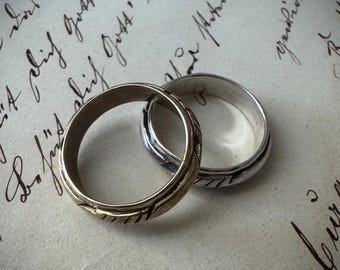 May include: Two wedding rings, one gold and one silver, rest on a vintage paper background with handwritten script. The gold ring has a textured design, while the silver ring is smooth. The rings are interlocked.