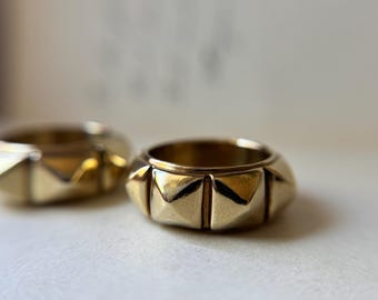 May include: Two gold-toned rings featuring a repeating pyramid stud pattern around the exterior band. The rings have a polished, reflective metallic finish with distinct geometric shapes forming a continuous textured design around the circumference.
