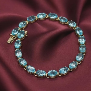 May include: A gold-toned bracelet featuring a series of oval-cut light blue gemstones linked together. The piece includes a secure metal clasp mechanism at one end, with each stone individually set in a polished gold-colored metal frame.