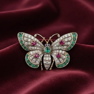May include: A detailed butterfly brooch with a gold-tone frame, encrusted with clear and red gemstones, and outlined with green gems. The butterfly's body features a central green gem. The brooch is set against a deep red fabric.