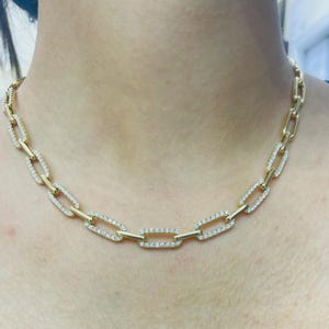 May include: A gold-colored chain necklace with rectangular links, each encrusted with small, sparkling white stones. The necklace is displayed around a person's neck, showcasing its design and detail.