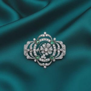 May include: An ornate, silver-toned brooch with a floral design. The brooch features numerous small, sparkling clear stones and square-cut green stones. The brooch is set against a teal-colored fabric background.