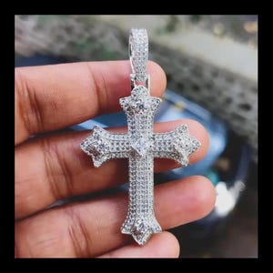 May include: A silver-colored cross pendant encrusted with numerous small, sparkling stones. The cross features a detailed design with pointed edges and a central, larger stone. The pendant is suspended from a matching bail.