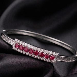 May include: A silver-toned hinged bangle bracelet featuring a central row of square-cut red rubies flanked by two rows of round brilliant-cut white diamonds, with additional baguette-cut white diamonds set along the sides of the metal band.