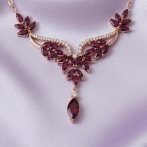 May include: A rose gold necklace featuring a central cluster of marquise-cut deep red garnet stones arranged in a leaf-like pattern, accented with small clear pavé crystals along the metal frame and a single dangling marquise-cut garnet drop.
