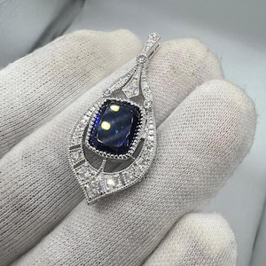 May include: A silver pendant with a large, rectangular, dark blue gemstone at its center. The pendant is surrounded by smaller, clear gemstones and intricate filigree detailing. The design is teardrop-shaped, with a loop at the top for a chain.