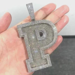 May include: A dazzling, silver-toned pendant in the shape of the letter "P", encrusted with numerous sparkling, clear gemstones. The pendant has a bail for a chain and is held in a hand, showcasing its intricate design and brilliance.