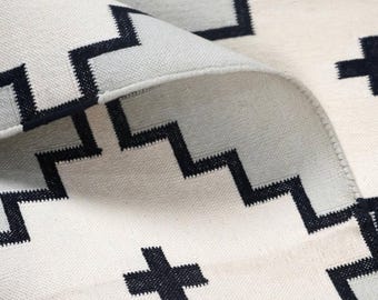 Puede incluir: Primer plano de una alfombra color crema con un patrón repetitivo de zigzags negros y signos de suma. La alfombra está doblada, revelando la parte inferior y el borde cosido. La alfombra parece estar hecha de un material tejido.