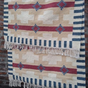 Op de afbeelding: Handgeweven tapijt met een geometrisch patroon. Het tapijt heeft een crèmekleurige achtergrond met rode en blauwe accenten. Het ontwerp omvat ruitvormen en strepen. De randen zijn afgewerkt met een blauwe rand en witte franjes.