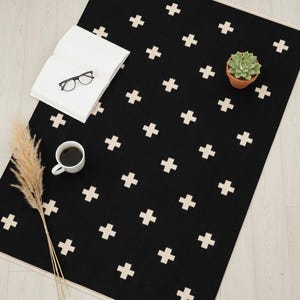 Può includere: Un tappeto nero con un motivo ripetuto di croci bianche. Il tappeto ha un bordo color crema. Un libro, occhiali, una piccola pianta in vaso, una tazza di caffè ed erba secca sono sul tappeto.