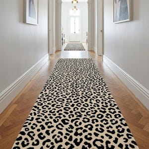 Könnte beinhalten: Ein langer, rechteckiger Läufer mit einem sich wiederholenden schwarzen Leopardenmuster auf cremefarbenem Grund. Der Teppich liegt mittig auf einem Holzboden in einem Flur und erstreckt sich vom Vordergrund bis zu einer entfernten Tür.