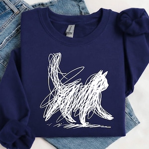 May include: A navy blue sweatshirt with a white, scribbled cat illustration. The cat is shown in profile, walking with its tail up. The sweatshirt is placed on top of a pair of blue denim jeans, creating a relaxed look.