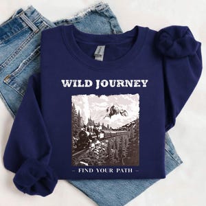 Könnte beinhalten: Marineblaues Sweatshirt mit dem Schriftzug "WILD JOURNEY" über einer Schwarz-Weiß-Grafik eines Zuges, der durch eine Berglandschaft fährt. Der Satz "FIND YOUR PATH" steht darunter. Das Sweatshirt liegt gefaltet auf einer Jeans.