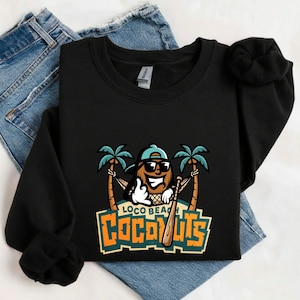 Op de afbeelding: Zwart sweatshirt met een afbeelding van een kokosnoot met zonnebril en een honkbalknuppel, met de tekst "LOCO BEACH COCONUTS" in oranje en wit. Het ontwerp bevat palmbomen en een hangmat. Het sweatshirt ligt op een blauwe jeans.