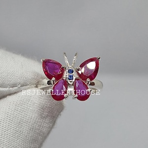 May include: A butterfly-shaped ring with ruby and sapphire gemstones. The ring features a silver band and a butterfly design with ruby teardrop-shaped wings and blue and black accents. The ring is a piece of jewelry.