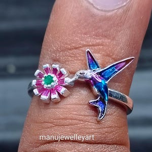 May include: A silver ring with a hummingbird and flower design. The hummingbird has blue and purple wings, and the flower has pink petals with a green centre. A distinctive piece of jewellery.