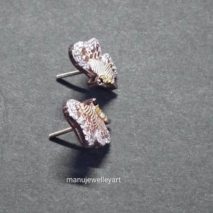 May include: A pair of butterfly stud earrings. The earrings feature a rose gold setting, with white and yellow detailing. The wings are embellished with small, clear crystals. They are presented on a dark grey surface.