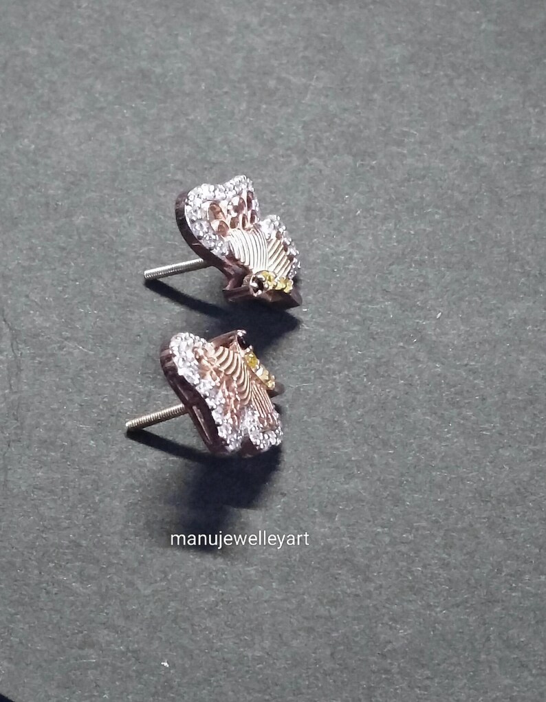 May include: A pair of butterfly stud earrings. The earrings feature a rose gold setting, with white and yellow detailing. The wings are embellished with small, clear crystals. They are presented on a dark grey surface.
