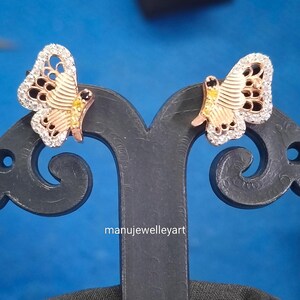 May include: A pair of butterfly earrings. The earrings have a rose gold body with white and black detailing. The wings are edged with small, clear stones. The earrings are displayed on a black stand against a blue backdrop.