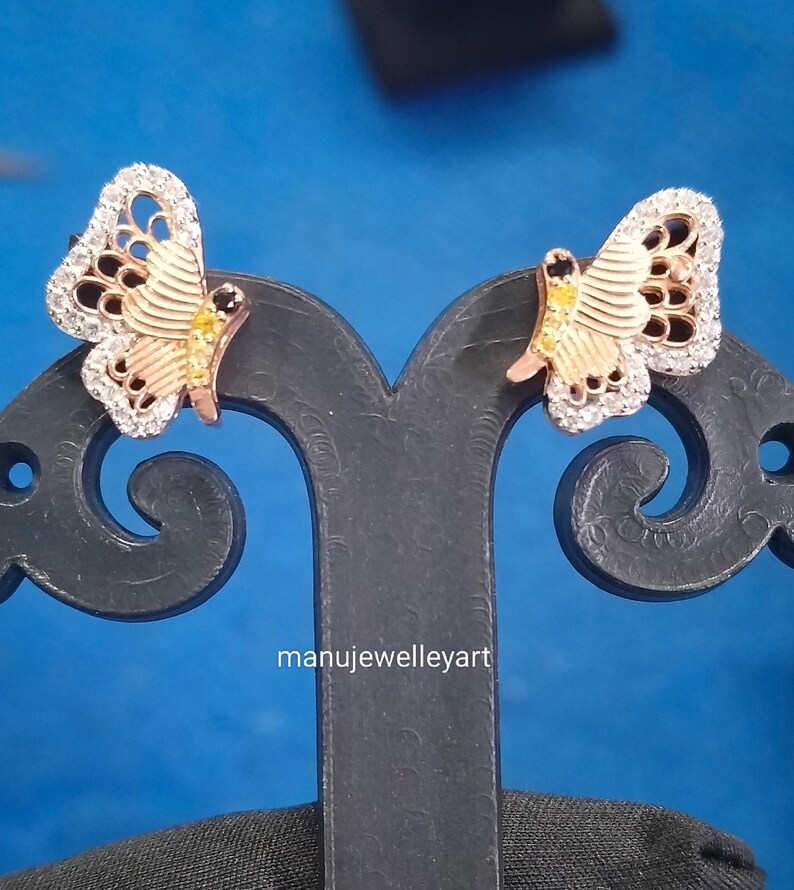 May include: A pair of butterfly earrings. The earrings have a rose gold body with white and black detailing. The wings are edged with small, clear stones. The earrings are displayed on a black stand against a blue backdrop.