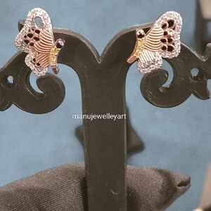 May include: A pair of butterfly earrings. The earrings have a silver outline, a rose gold body, and black detailing. The wings are set with small, clear stones. The earrings are displayed on a black stand. The text "manujewelleyart" is visible.