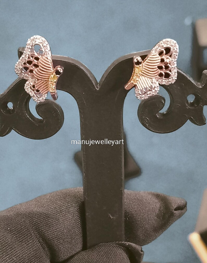 May include: A pair of butterfly earrings. The earrings have a silver outline, a rose gold body, and black detailing. The wings are set with small, clear stones. The earrings are displayed on a black stand. The text "manujewelleyart" is visible.