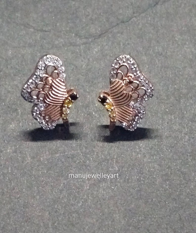 May include: A pair of butterfly earrings. The earrings have a gold-coloured body with white and yellow gemstones. The wings are outlined with white gemstones, and the body has a striped design. The earrings are set against a grey background.