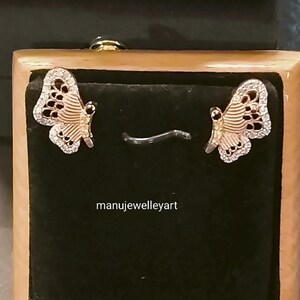 May include: Butterfly earrings presented in a wooden jewellery box. The earrings have a gold-coloured body and wings adorned with small, clear gemstones. The wings have a detailed pattern with black accents. The text "manujewelleryart" is visible.