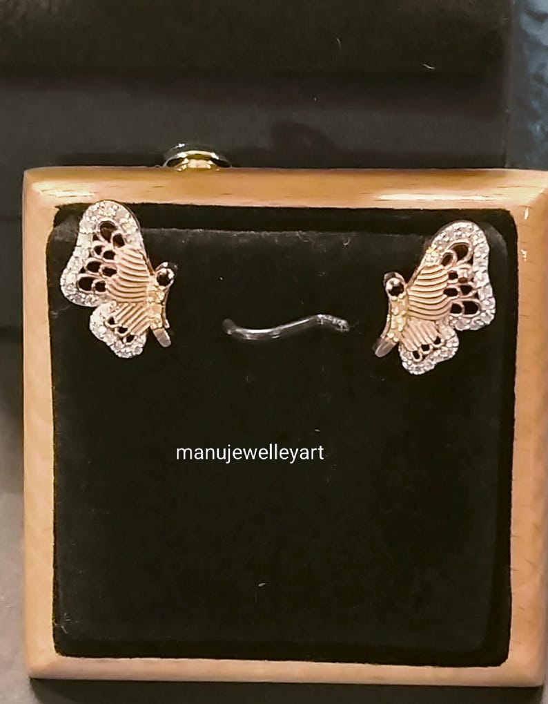 May include: Butterfly earrings presented in a wooden jewellery box. The earrings have a gold-coloured body and wings adorned with small, clear gemstones. The wings have a detailed pattern with black accents. The text "manujewelleryart" is visible.