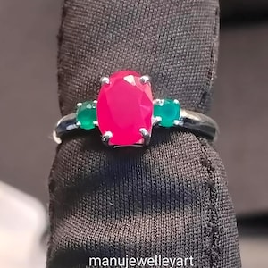 Puede incluir: Un anillo de plata con una gran gema rosa brillante de talla ovalada flanqueada por dos gemas verdes redondas más pequeñas. El anillo tiene una banda fina y pulida. El texto "manujewelleyart" es visible.