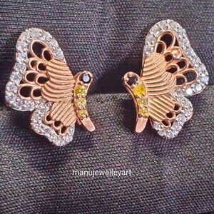 May include: A pair of rose gold butterfly earrings. The wings are detailed with white gemstones and the body features yellow and black gemstones. The earrings are displayed on a dark grey surface. The text "manujewelleyart" is visible.