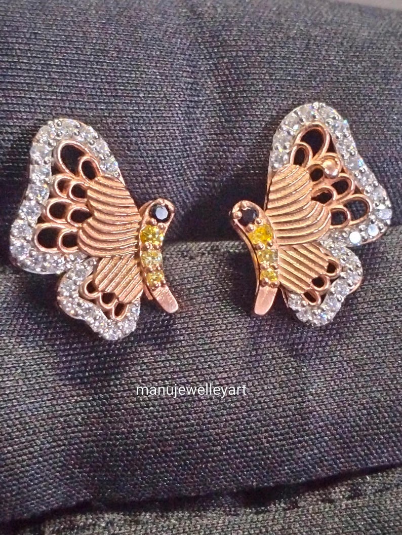 May include: A pair of rose gold butterfly earrings. The wings are detailed with white gemstones and the body features yellow and black gemstones. The earrings are displayed on a dark grey surface. The text "manujewelleyart" is visible.