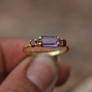 Op de afbeelding: Een gouden ring met een rechthoekige paarse edelsteen geflankeerd door twee kleinere rode edelstenen. De ring heeft een delicate band en de tekst "RKHSILVERGEMS" is zichtbaar.
