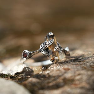 Puede incluir: Anillo de plata con una gema rectangular de color ámbar. El anillo tiene un diseño de hojas y vides con una pequeña gema roja redonda. El texto "RKHSILVERGEMS" es visible en el anillo.
