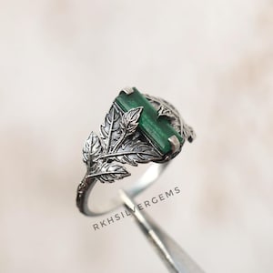 May include: A silver ring with a rectangular, emerald-green gemstone. The ring has a detailed leaf design on the sides and around the stone. The text "RKHSILVERGEMS" is visible on the ring.