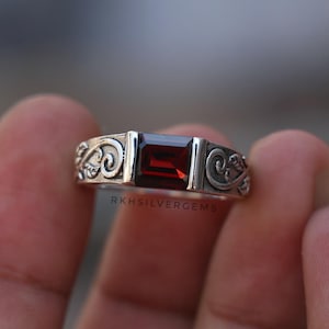 May include: A silver ring with a rectangular, deep red gemstone. The band has an ornate, swirling pattern. The text "RKHSILVERGEMS" is visible. The image is a close-up, showing the ring's intricate details.